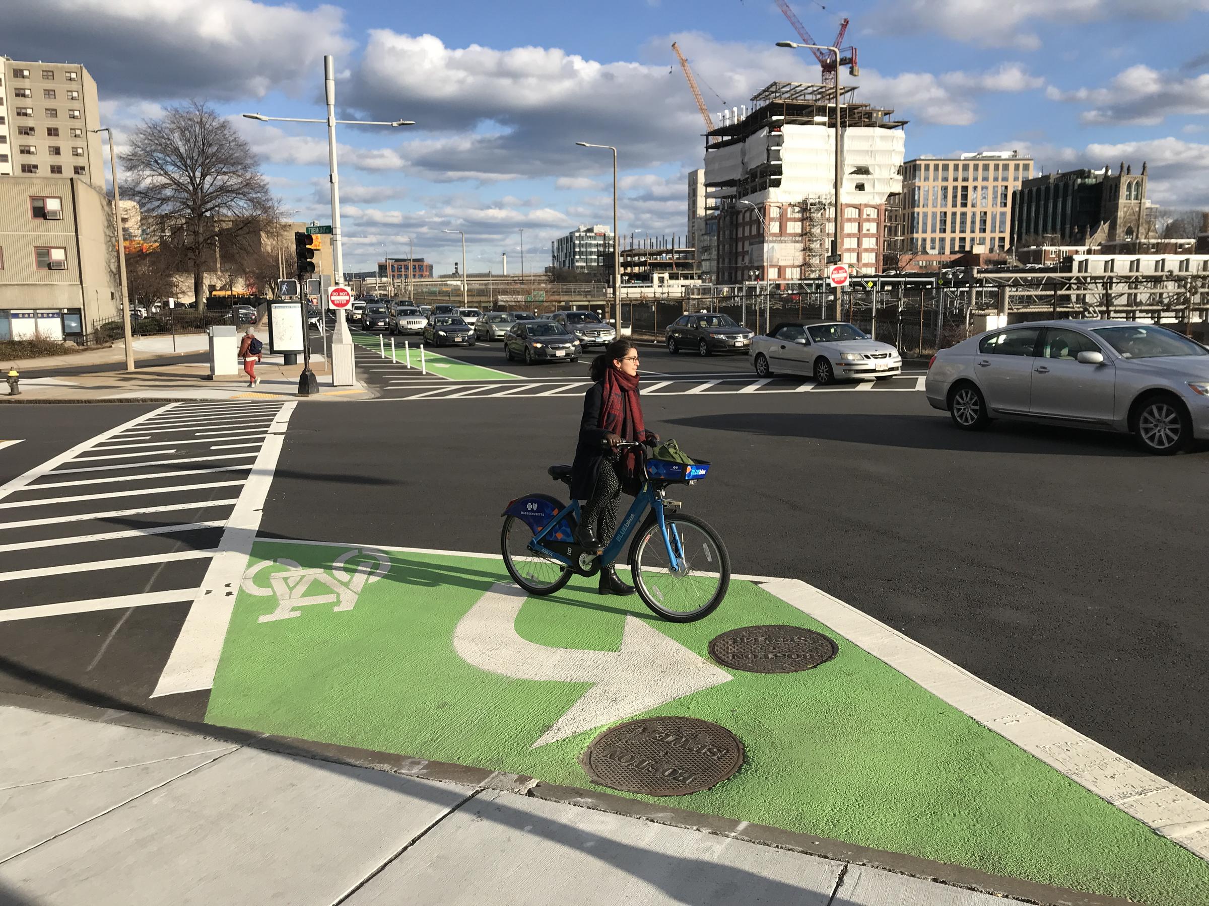 Photo showing a two-stage turn box. In the foreground, the two-stage turn box is visible. A bicyclist is waiting in the box to proceed. In the background, a bike lane is visible on the perpendicular street.