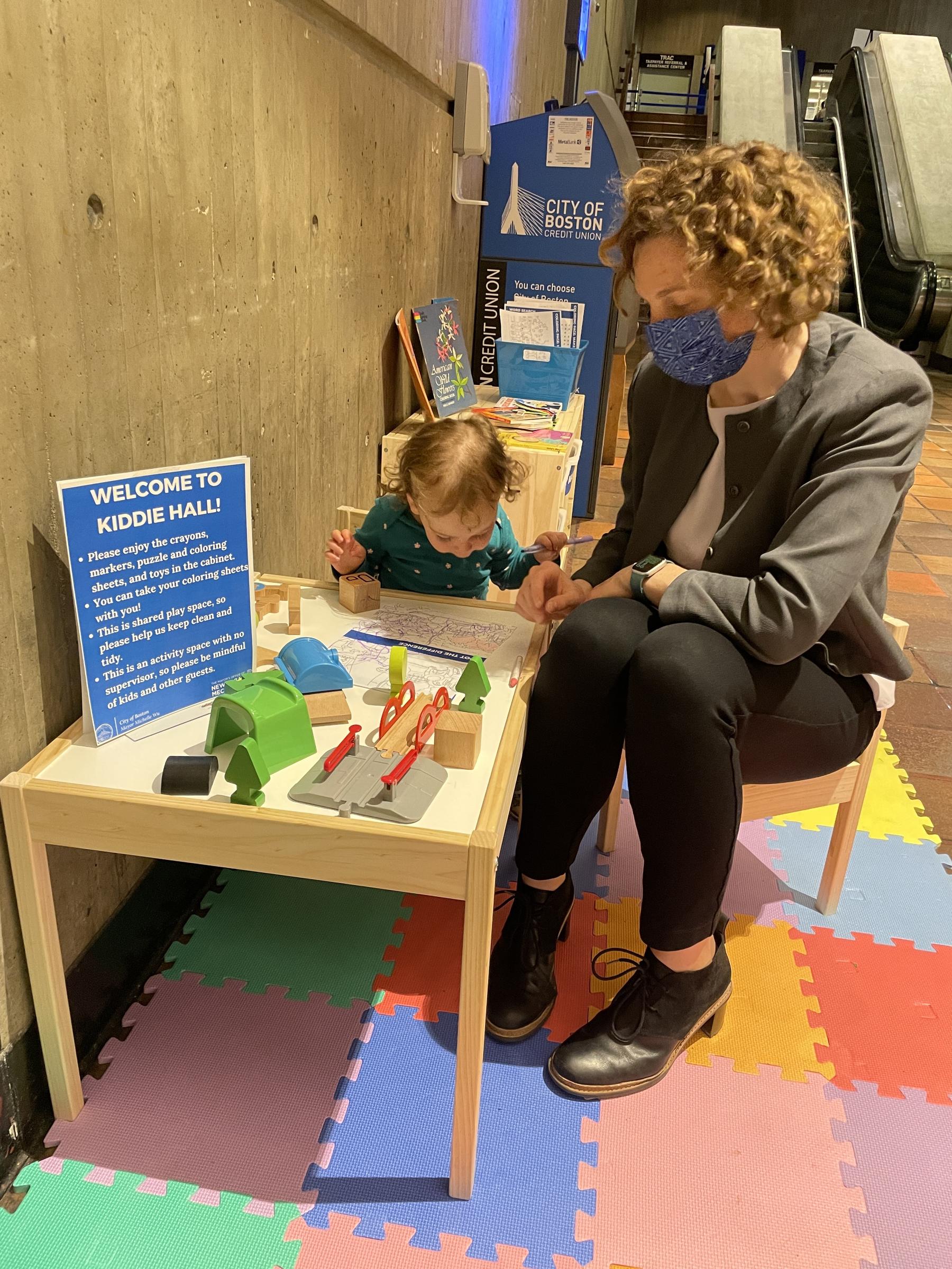 A child and an adult sit at a table on colorful tiles in front of a concrete wall