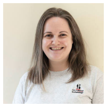 A smiling woman with light brown hair in front of a beige wall. She is wearing a grey t-shirt with the City of Boston Archaeology Program logo .