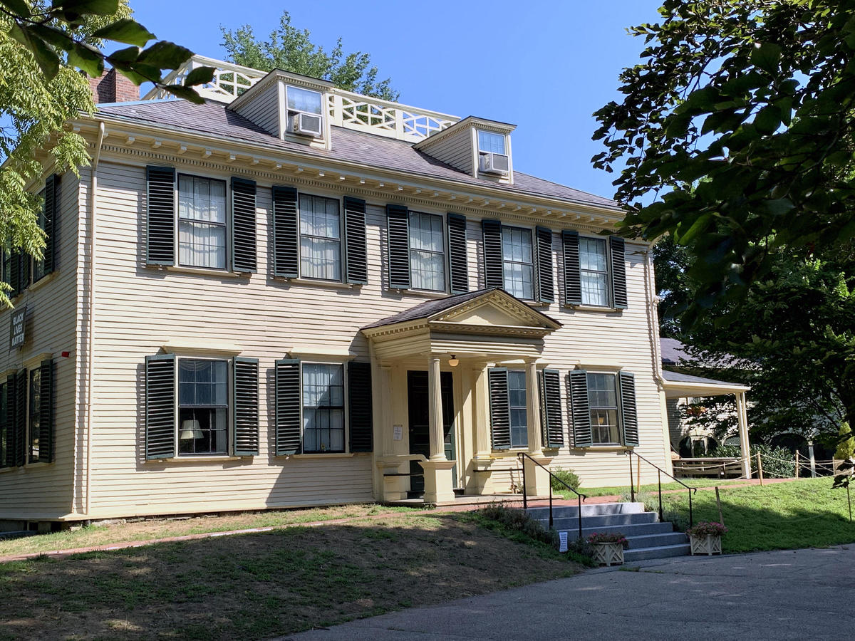 View of an 18th-century Georgian mansion with light yellow siding and dark green shutters. An ell extends off to the right.