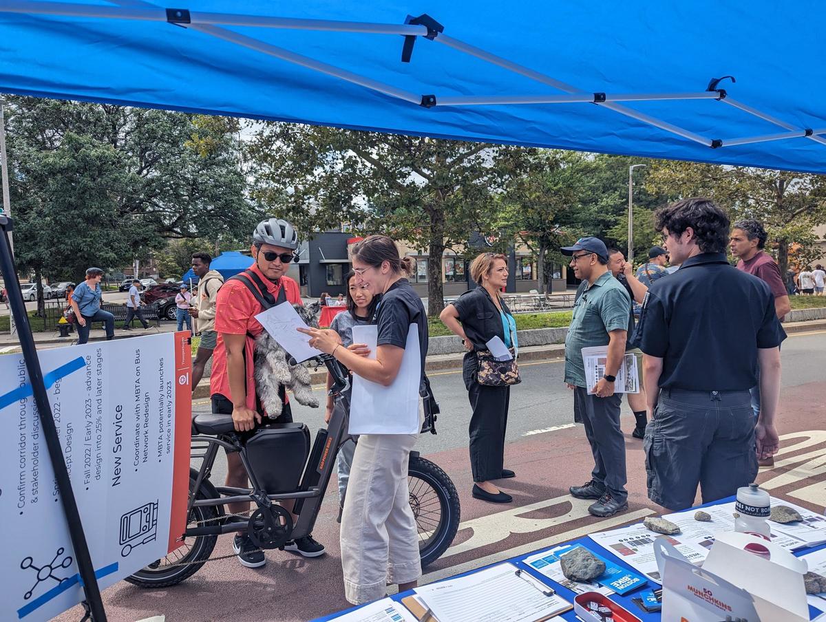 A BTD planner talks to constituents about transportation projects comming to Allston Brighton. They stand near a blue tent and a bicycle.