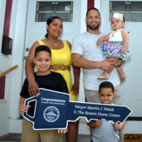 Image of family standing in front of housing, holding a symbolic "key" from the Boston Home Center.