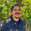 A picture of a person in front of a tree. The person has some facial hair and is wearing a blue shirt.