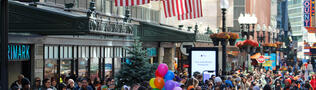 A large crowd of people on Summer Street under American flags. A woman holds balloons at the center.